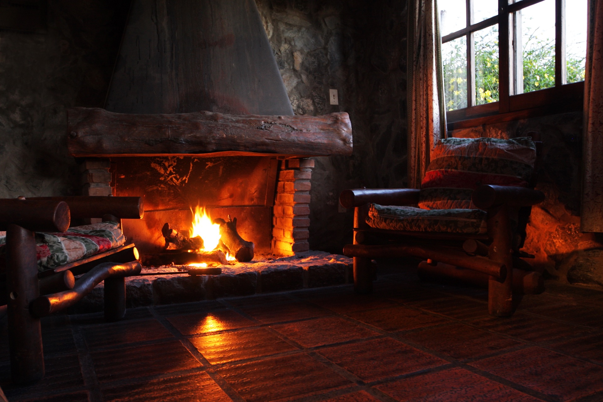 The stone hearth blazing — log chairs pulled close, evening light in the window